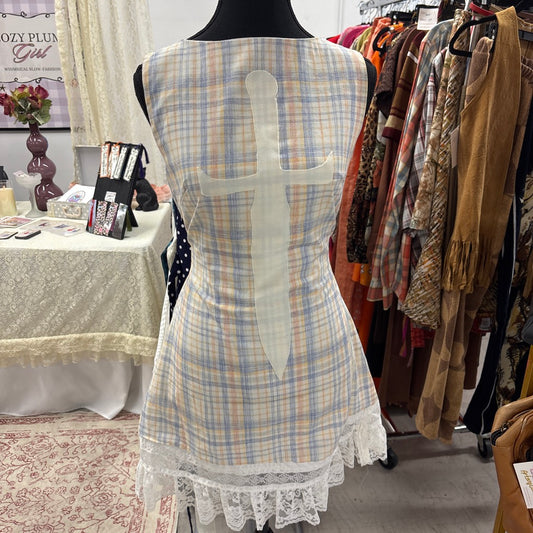 A sleeveless plaid dress displayed on a black mannequin torso in a retail setting. The dress has a pastel plaid pattern with blue, yellow, and white tones, a large white cross-shaped cutout on the back, and white lace trim at the hem.